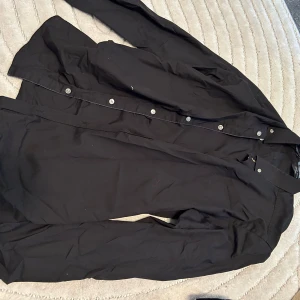 Svart skjorta med knappar - Snygg svart skjorta med klassisk krage och silverfärgade knappar längs hela framsidan. Skjortan har lång ärm och är tillverkad i ett mjukt och lätt material som känns skönt mot huden. Perfekt för dig som gillar stilrena och enkla plagg.