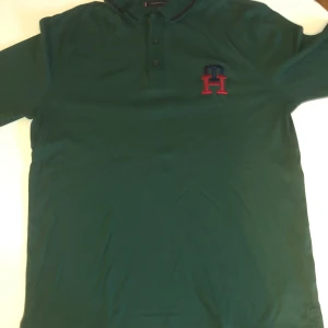 Grön Tommy Hilfiger långärmad tröja slim fit - Snygg grön långärmad tröja från Tommy Hilfiger i slim fit med korta ärmar. Har broderad TH-logga i blått och rött på bröstet och klassisk Hilfiger-flagga på ärmen. Mjuk bomullskvalitet och knappslå vid halsen för en clean look.