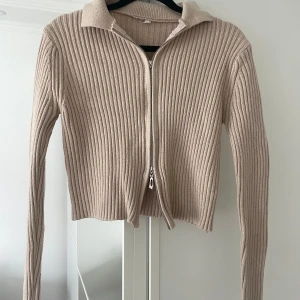 Beige ribbad kofta med dragkedja - Säljer en beige ribbad kofta med lång ärm och krage. Tröjan har en hel dragkedja framtill och är croppad i modellen.