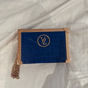 Blå kuvertväska från Yves Lansac -  blå kuvertväska från Yves Lansac med krokopräglat mönster och guldfärgade metalldetaljer. Väskan har en kedjerem i guld och en boxig form.