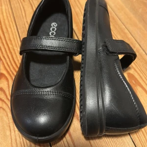 Svarta ballerinaskor från ECCO i skinn - Stilrena svarta ballerinaskor från ECCO i mjukt skinn med klassisk rund tå och praktiskt kardborreband över foten. Skorna har platt sula och diskreta sömdetaljer framtill. Perfekta för en clean och enkel look.