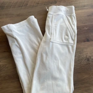 Vita mjukisbyxor från Juicy Couture - Snygga vita mjukisbyxor från Juicy Couture med broderad logga på fickan. Byxorna har snörning i midjan, breda ben och en ficka med knapp på baksidan. Mjukt och skönt material som är perfekt för chill dagar.