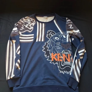Blå Kenzo tröja med tigertryck - Säljer en blå långärmad tröja från Kenzo med stort tigertryck och logga i orange framtill. Tröjan har coola vita och gråa ränder på ärmar och axlar, samt rund hals. Perfekt för dig som gillar statement-plagg och streetstyle.