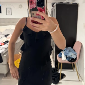 Svart volangklänning med smala axelband - Säljer en svart kortklänning med smala axelband och snygga volangdetaljer framtill och på sidorna. Klänningen har en figurnära passform och är perfekt för dig som gillar en stilren look med lite extra detaljer. Superfin att styla med klackar eller sneakers.