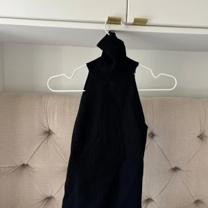 Svart ribbad ärmlös klänning - Snygg svart topp med ribbad struktur och hög hals. Modellen är ärmlös och har en tight passform som framhäver siluetten. Perfekt för dig som gillar minimalistisk stil och vill ha något enkelt men ändå trendigt.