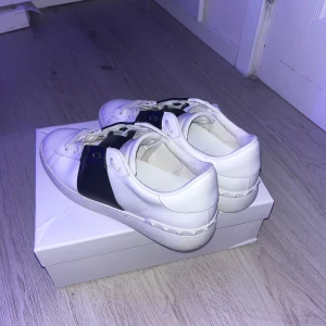 Valentino Garavani Open sneakers vit/svart - Snygga Valentino Garavani Open sneakers i vitt skinn med bred svart läderdetalj över vristen. Klassisk rund tå, vita snören och ikoniska nitar på hälen. Perfekta för dig som vill ha en lyxig och stilren look.