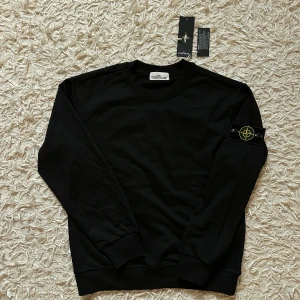 Stone Island- svart sweatshirt med lapparna kvar  - Säljer en helt ny Stone island sweater i den klassiska svarta färgen. Denna svarta Stone island tröja har det ikoniska kompass märket på vänstra arm och är tillverkad av mjukt, bekväm material, perfekt för avslappnad vardagsstil. Nyskick. Denna har aldrig burits och har alla original lappar kvar. Storlek M.  