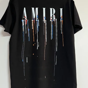 Svart AMIRI t-shirt med färgstänk - Svart t-shirt från AMIRI med stort tryck av loggan och färgstänk i blått, vitt, rött och orange på både fram- och baksida. Klassisk passform med rund hals och korta ärmar. Materialet är mjuk bomull som känns skön mot huden. Perfekt för dig som gillar streetwear med edge.