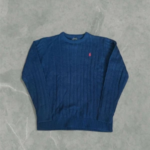 Marinblå kabelstickad tröja från Polo Ralph Lauren - Kabelstickad blå tröja från Polo Ralph Lauren med rund hals och klassisk röd broderad logga på bröstet. Det är storlek L och tröjan är i mycket bra skick