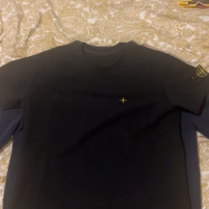 Svart t-shirt från Stone Island - Svart t-shirt från Stone Island med klassisk rund hals och korta ärmar. På bröstet finns en liten gul broderad detalj och på vänster ärm sitter den ikoniska Stone Island-loggan i gult. Perfekt för dig som gillar stilrena och exklusiva plagg. Skriv för fler bilder eller funderingar.