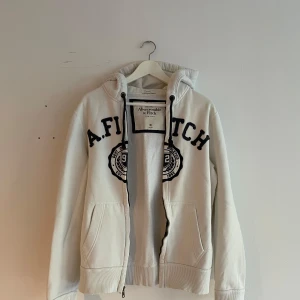Vit hoodie från Abercrombie & Fitch - Vit hoodie från Abercrombie & Fitch med svart broderad logga och emblem på framsidan. Tröjan har huva med snörning, ribbade muddar och fickor fram. Materialet är mjuk bomull och polyester, perfekt för chill dagar. Dragkedja framtill för enkel på- och avtagning.