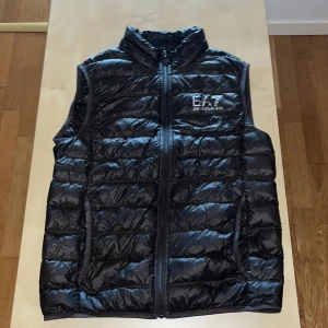Mörkgrå dunväst från Emporio Armani EA7 - Snygg mörkgrå dunväst från Emporio Armani EA7 med quiltad design och hög krage. Västen har dragkedja framtill och två fickor på sidorna. Perfekt att slänga över en hoodie för en sportig look. EA7-logga på bröstet ger en exklusiv touch.