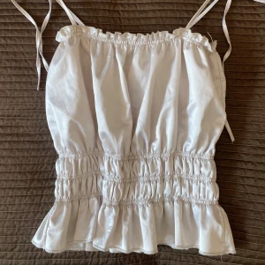 Ljus silkestopp med knytband - Supersöt topp i ljus färg med smala axelband som knyts på axlarna. Toppen har volangkant nertill och flera rader med rynk för en puffig och luftig look. 