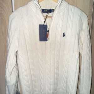Vit stickad tröja från Polo Ralph Lauren - Säljer en vit kabelstickad tröja från Polo Ralph Lauren med half zip och ribbad krage. Tröjan har långärmad passform och klassisk broderad logga på bröstet. Perfekt för dig som gillar stilrena och tidlösa plagg med snygga detaljer.