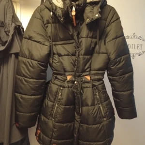 Svart pufferjacka från Navahoo - Snygg svart pufferjacka från Navahoo med huva och mysigt grått teddyfoder. Jackan har dragkedja, snörning i midjan och flera fickor med detaljer i brunt. Perfekt för kalla dagar och har en cool badge på ryggen.