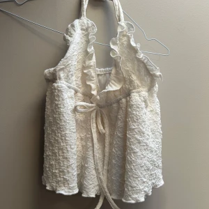 Vit volangtop med knyt och struktur - Supersöt vit linnetopp med volangkant längs urringningen och knyt i midjan. Toppen har en strukturerad, krinklad yta och är ärmlös med halterneck. Perfekt för varma dagar när du vill känna dig extra fin.