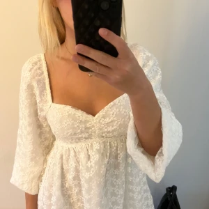 Vit spetsklänning med puffärmar - Superfin vit kort klänning med romantisk spets och blommigt mönster. Klänningen har puffiga långärmar, söt v-ringning och markerad midja. Perfekt för sommarens alla tillfällen och ger en dreamy vibe. Helt ny med lapp kvar!