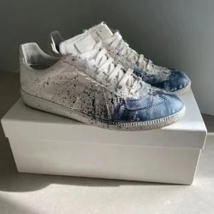 maison margiela skor - Unika sneakers i vitt och blått med coola färgstänk och slitna detaljer. Skorna har snörning och en platt sula. Ovandelen är i canvas och de har en rund tå. Perfekta för dig som gillar en kreativ och edgy stil.