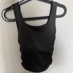 Svart topp - Säljer en stilren svart topp med breda axelband och rynkade sidor som ger en snygg passform. Klassisk modell med rundad halsringning både fram och bak. Oanvänd 