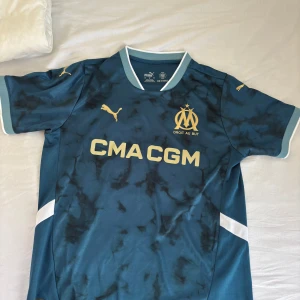 Marinblå Marseille fotbollströja Puma - Snygg marinblå Olympique de Marseille fotbollströja från Puma med gul logga och tryck. Tröjan har korta ärmar, vit krage och detaljer samt ett unikt mönster över axlarna. Materialet är lätt och andas, perfekt för match eller träning.