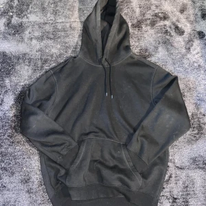 Svart hoodie med huva och ficka - Oversized Svart hoodie med klassisk huva och snörning, samt stor magficka framtill. Tröjan har långärmade muddar och en loose passform som är chill och enkel att bära. Perfekt för att slänga på sig när det är lite kallare ute.