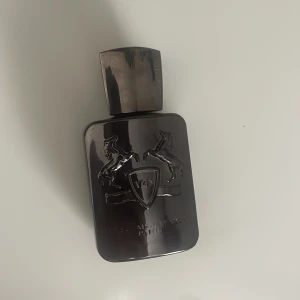 Parfym Parfums de Marly Herod - Snygg parfymflaska i metallisk mörkgrå färg från Parfums de Marly, modell Herod. Flaskan har en stilren, fyrkantig form med ingraverade hästar och sköld på framsidan samt ett robust lock. Perfekt för dig som gillar lyxiga och unika dofter.