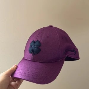 Lila keps från Black Clover - Säljer en snygg lila keps från Black Clover med svart fyrklöver framtill och broderad texten 'Live Lucky' på sidan. Kepsen har böjd skärm och är tillverkad i ett mjukt syntetmaterial. Storlek S/M. Passar för både tjej och kille. Oanvänd.