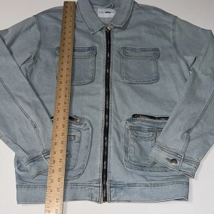 Ljusblå jeansjacka från Fashion Nova - Snygg ljusblå jeansjacka från Fashion Nova med fyra fickor framtill, två med dragkedja och två med lock. Jackan har klassisk krage, långa ärmar med knapp vid ärmslut och hel dragkedja framtill. Perfekt för dig som gillar streetstyle och denim.