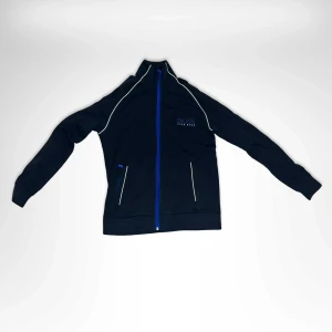 Svart ziptröja från BOSS med blå detaljer - Svart långärmad ziptröja från BOSS med blå dragkedja och logga på bröstet. Snygga vita kontrastlinjer längs axlar och fickor. Tröjan har hög krage och är tillverkad i mjuk bomullsmix. Perfekt för en sportig och clean look.