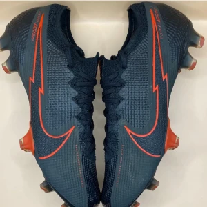 Nike Mercurial Vapor 13 fotbollsskor - Svarta Nike Mercurial Vapor 360 fotbollsskor med röda detaljer och stora Swoosh-loggor. Skorna har snörning, stickad ovandel i syntetmaterial och mönstrad struktur för extra grepp. Sulan har röda och svarta dobbar för bästa fäste på planen.