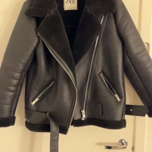 Svart skinnjacka från Zara - Snygg svart skinnjacka från Zara med mjukt foder och stiliga dragkedjedetaljer. Jackan har en klassisk krage och ett bälte i midjan för extra stil. Perfekt för kyligare dagar.