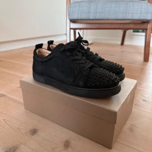 Svarta sneakers med nitar från Christian Louboutin - Snygga svarta sneakers från Christian Louboutin med nitar på ovansidan och den ikoniska röda sulan. Skorna har snörning och är tillverkade i skinn, vilket ger en lyxig känsla. Perfekta för den som vill sticka ut med stil.
