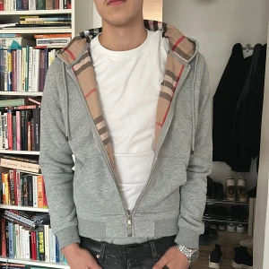 Burberry Hoodie - Säljer en stilren grå hoodie från Burberry med det klassiska rutiga mönstret på insidan av huvan. Den har dragkedja framtill och är tillverkad i mjukt material för extra komfort. Perfekt för en avslappnad look.