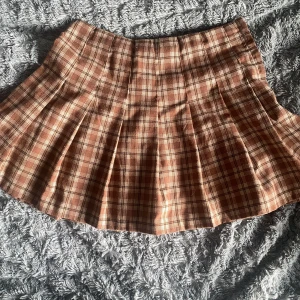 Brun rutig veckad kjol - Brun rutig kortkjol med veckad design. Kjolen har en klassisk rutig stil i bruna och beige toner. Ca 38cm lång och 35cm i midjan