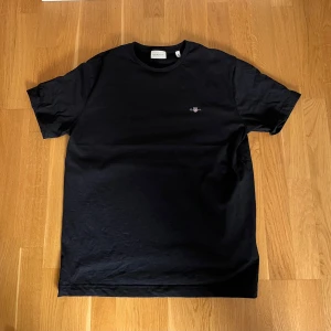 Gant T-Shirt  - Svart GANT T-shirt 👕 i storlek M – stilren och klassisk! Tillverkad av 100% bomull ☁️ för maximal komfort. Perfekt för både vardag och avslappnade tillfällen! Diskret logga på bröstet för en clean look ✨                                                                                    Aldrig använd, bara utan prislapp!  