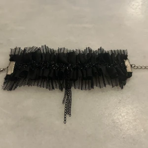 Svart spetschoker med volang armband - Elegant svart choker med volangdetaljer och spets. Kedja i silverfärg för justerbar passform. Perfekt för att ge en dramatisk touch till din outfit.