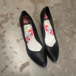 Svarta pumps  - Eleganta svarta pumps från Vagabond med spetsig tå och klack. Insidan har ett färgglatt blommönster i rött och gult. Perfekta för en stilren look.