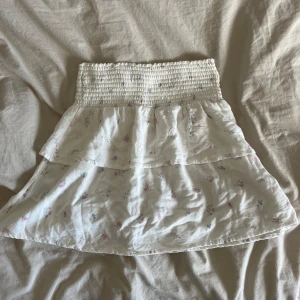 Vit blommig kjol - Söt vit kjol med blommigt mönster och volangdetaljer. Den har en elastisk midja för en bekväm passform. Perfekt för varma dagar! Storlek 160 men passar xs