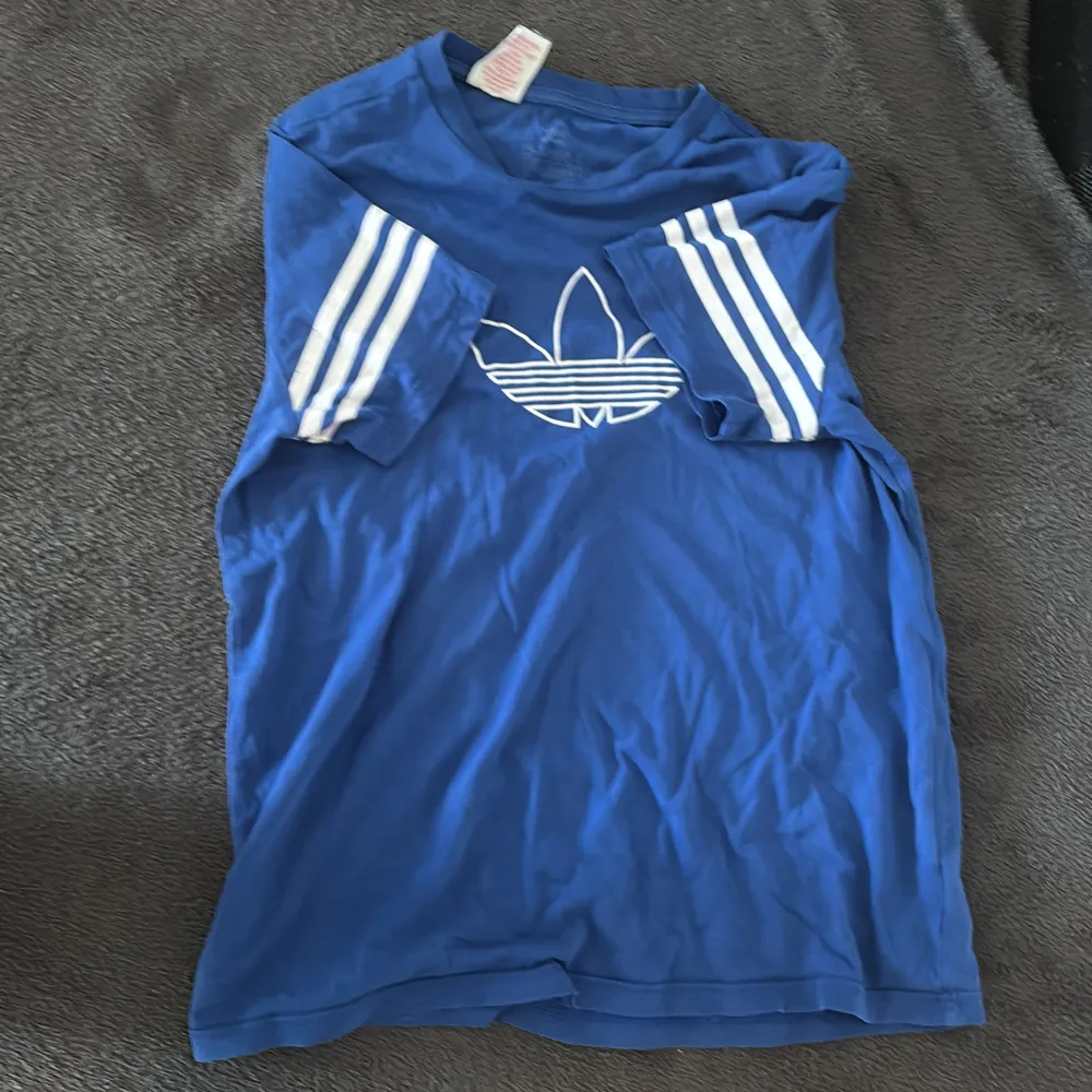 Säljer en blå t-shirt från Adidas med det klassiska trefoil-logotypen tryckt på framsidan. T-shirten har korta ärmar med vita ränder och är perfekt för en sportig look. Tillverkad i mjukt material för komfort hela dagen.. T-paidat.
