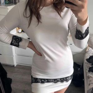 Vit klänning med spets - Snygg vit klänning med svarta spetsdetaljer vid ärmslut och nederkant. Klänningen har långa ärmar och en tight passform. (Katt och kanin finns i hemmet) 