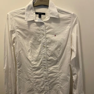 Vit skjorta från GANT - (Storlek 158-164) Säljer en stilren vit skjorta från GANT med broderad logotyp på bröstet. Skjortan har långa ärmar och knappar framtill. Perfekt för en klassisk look.