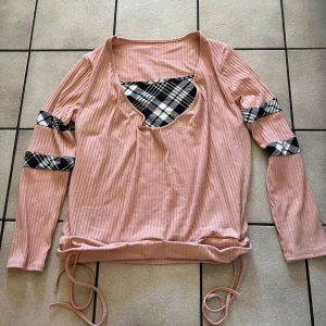 Rosa ribbad tröja med rutiga detaljer storlek 2XL - Säljer en rosa ribbad tröja med unika rutiga detaljer i svart och vitt på ärmarna och bröstet. Tröjan har långa ärmar och en knytning i nederkanten för justerbar passform. Perfekt för en avslappnad stil.