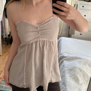 Topp med spetsdetaljer - Säljer ett sött linne med smala axelband och spetsdetaljer. Toppen har en vacker struktur och små knappar framtill. Perfekt för varma dagar eller som ett lager under en kofta.