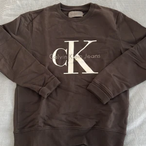 Brun sweatshirt från Calvin Klein Jeans - Säljer en stilren brun sweatshirt från Calvin Klein Jeans med det ikoniska CK-logotypen i vitt på framsidan. Tröjan har långa ärmar och ribbade muddar vid ärmslut och nederkant. Perfekt för en avslappnad look.