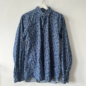 Blå blommig skjorta från John Henric - Snygg blå skjorta från John Henric med ett stilrent blommigt mönster i vitt. Skjortan har långa ärmar och knappar framtill. Perfekt för en elegant look.