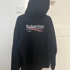 Svart hoodie från Balenciaga - Storlek M. Säljer en svart hoodie från Balenciaga med tryck både fram och bak. Hoodien har en klassisk design med dragsko i huvan och en stor ficka framtill. Perfekt för en stilren och avslappnad look.