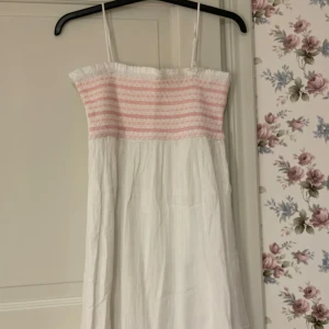 Vit och rosa klänning med smala axelband - Charmig vit klänning med rosa detaljer och smala axelband. Den har en elastisk överdel med rosa ränder och en luftig kjol. Perfekt för varma dagar! Aldrig använd!