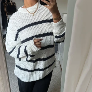 Vit och svart randig stickad tröja - Säljer en mysig vit stickad tröja med svarta ränder. Tröjan har en avslappnad passform och långa ärmar, perfekt för kyligare dagar. Den ribbade strukturen ger en extra touch av stil. Perfekt för en casual look!