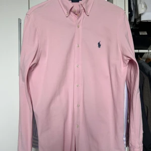 Rosa skjorta från Ralph Lauren - Säljer en stilren rosa knited Oxford slim fit  skjorta från Ralph Lauren med klassisk button down krage och knappar framtill. Enfärgad broderad logo på bröstet. Använd sparsamt utan tecken på slitage. 
