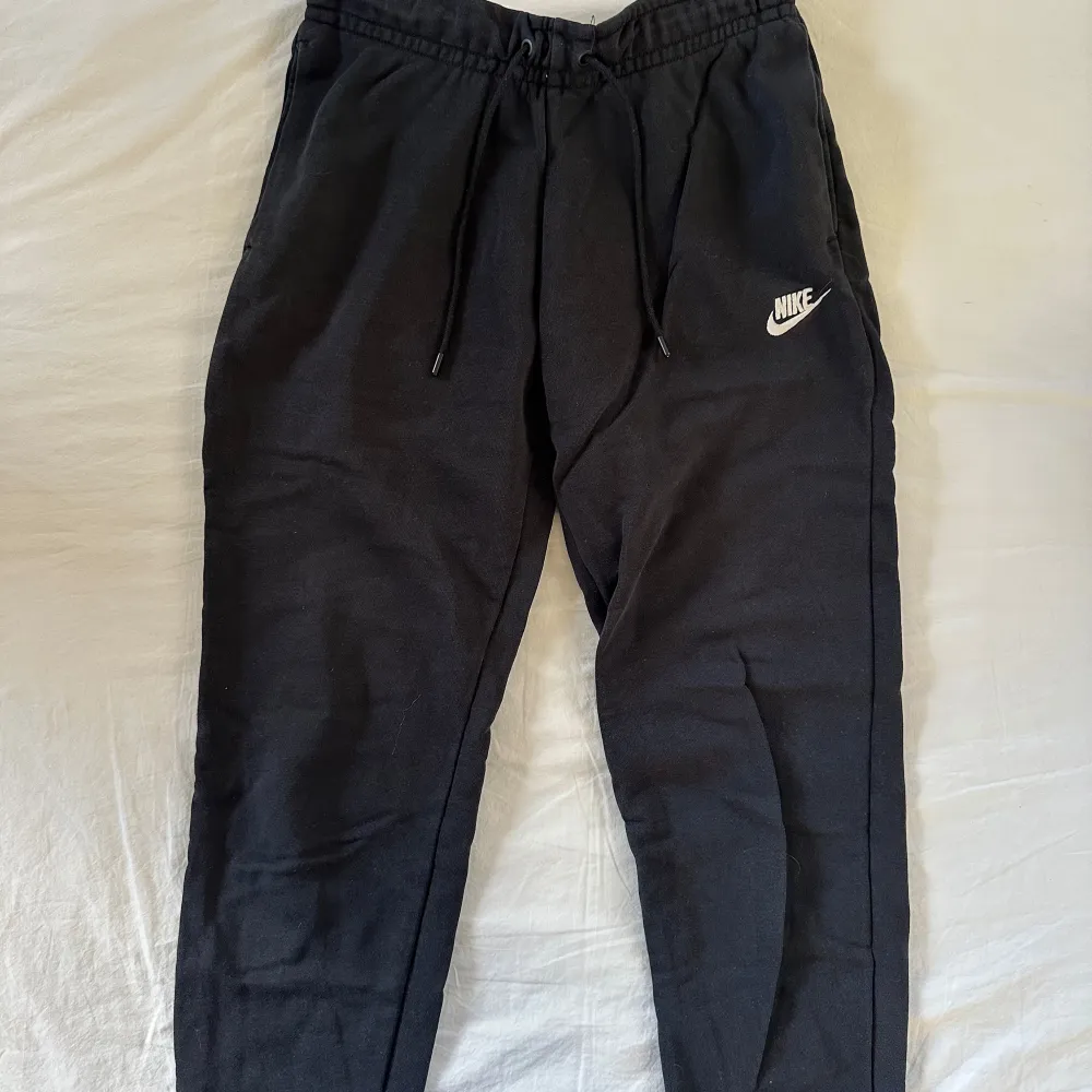Säljer ett par svarta mjukisbyxor från Nike med elastisk midja och snörning. Byxorna har en liten vit Nike-logga på benet och är perfekta för en avslappnad stil. Tillverkade i mjukt material för extra komfort.. Farkut & Housut.